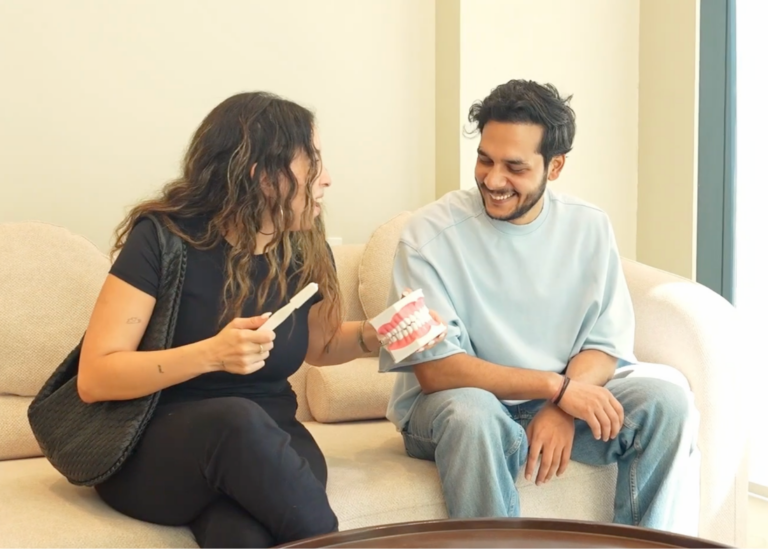 Woman shows a toothbrush to a smiling man while discussing a dental model on a beige sofa.