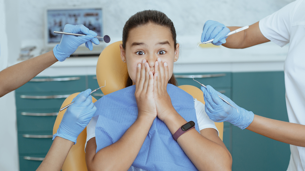 Girl in a dental chair covers her mouth as multiple dentists in blue gloves work around her with dental tools nearby behind at a clinic.
