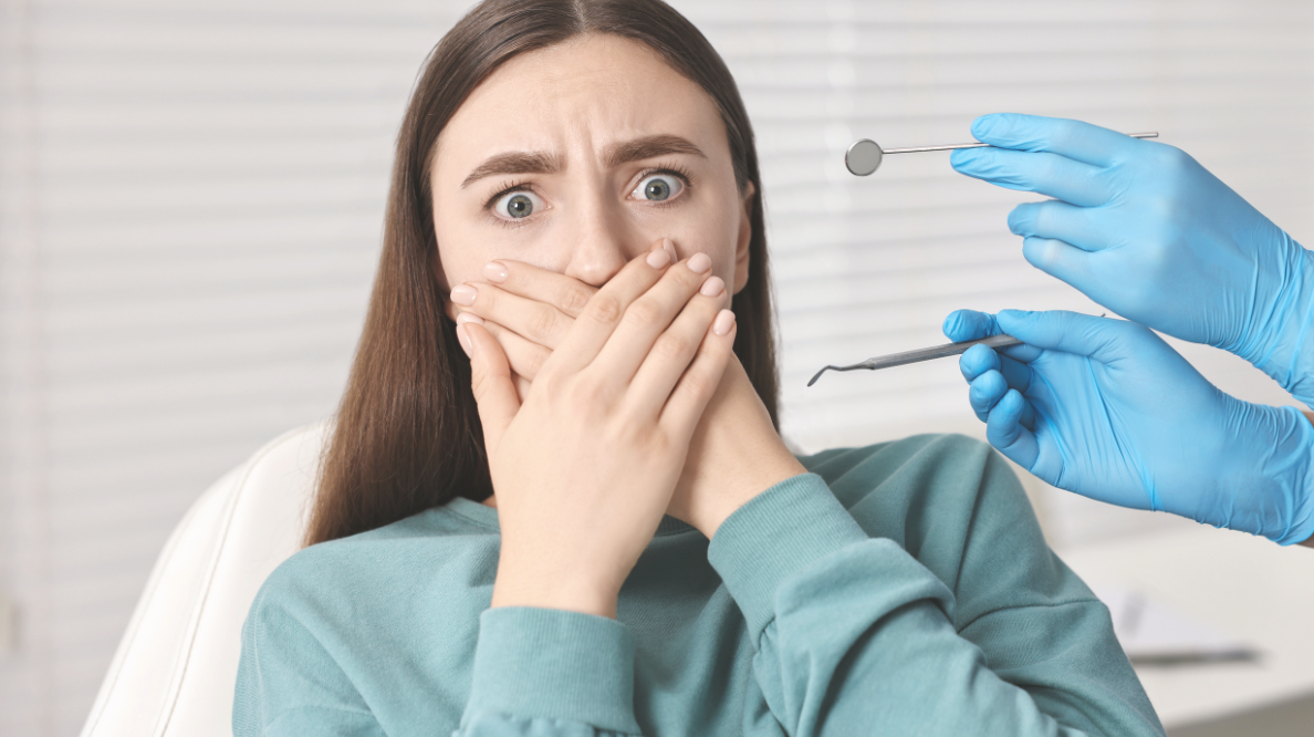 Woman seated in a dental chair covers her mouth in fear as a dentist in blue gloves holds a mirror and a dental instrument nearby.