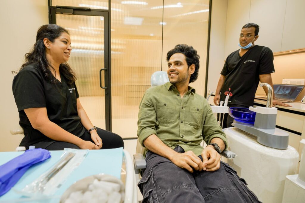Man in green shirt smiling while seated in a dental chair, conversing with a female clinician in black scrubs beside him in a clinic room with glass door behind.