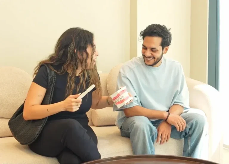 Consultation in the clinic lounge - Dr. Spandanas Dentistry - Dr. Spandanas Dentistry Woman and man sit on a beige sofa, smiling as the woman shows a toothbrush and a dental model with braces to explain dental care.