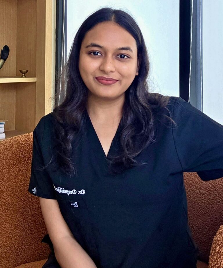 Woman in black medical scrubs sitting on a brown textured couch by a window, smiling at the camera, in a relaxed setting.