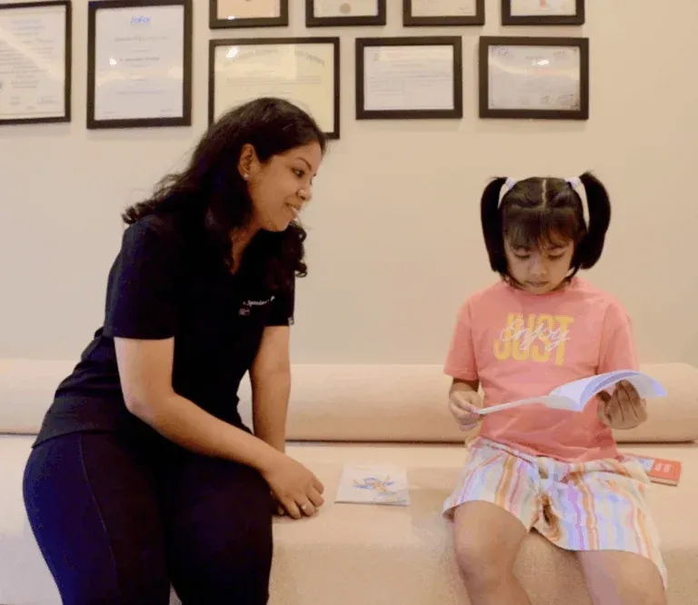 dr-spandanas-dentistry-pediatric-dentist-near-me-768x666 - Dr. Spandanas Dentistry - Dr. Spandanas Dentistry Woman kneels beside a seated girl on a beige sofa, reading a book together; framed certificates on the wall behind them.